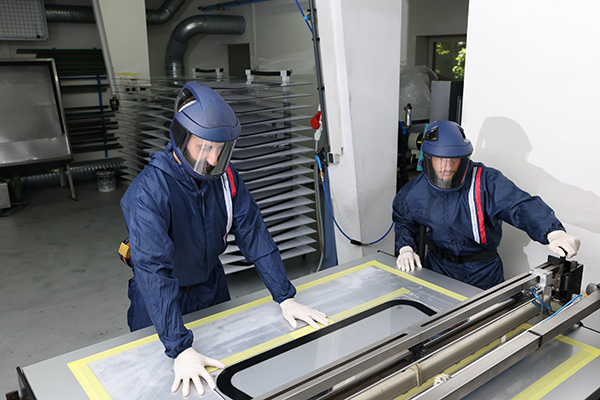 Zwei Männer in Schutzanzügen, die ein Kunststoffbauteil im Siebdruckverfahren bedrucken.